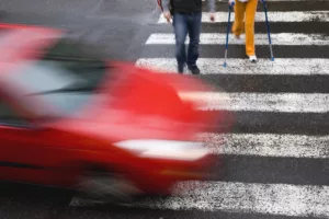 
A Blurred Red Car Speeds Through A Crosswalk Where Pedestrians—Including One On Crutches—Are Crossing, A Scene An Arlington Pedestrian Accident Lawyer Might Investigate.