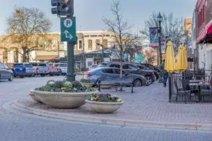 A Lively Street Scene In Mckinney, Texas, Featuring Cars Parked Along A Charming Sidewalk Cafe Area With Colorful Flower Pots, Reflecting The Vibrant Community That A Mckinney Personal Injury Lawyer Serves.