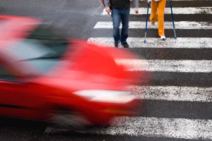 Red Car Blurring Through A Crosswalk With Pedestrians Stepping Into The Street, Representing The Urgency Of Contacting A Dallas Pedestrian Accident Lawyer After A Collision.