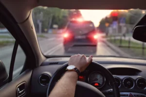 A Driver’s Perspective Inside A Car, Showing A Hand On The Steering Wheel With A Watch, While The Vehicle Is Stopped Behind Another Car With Illuminated Brake Lights At A Red Light. The Scene Prompts The Question: Is Brake Checking Illegal?