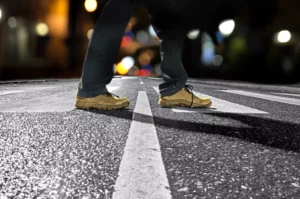 A Pedestrian’s Boots Cross Wet, Reflective Crosswalk Lines At Night While A Cockrell Hill Pedestrian Accident Lawyer Evaluates The Scene.