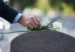Hand Placing A White Rose On A Gravestone, Symbolizing Grief And Loss—Representing The Emotional Impact Addressed By An Irving Wrongful Death Lawyer.