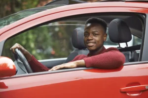 Teenage Driver Smiling While Wearing A Seatbelt And Sitting Behind The Wheel Of A Red Car, Illustrating Teenage Driver Safety Tips.